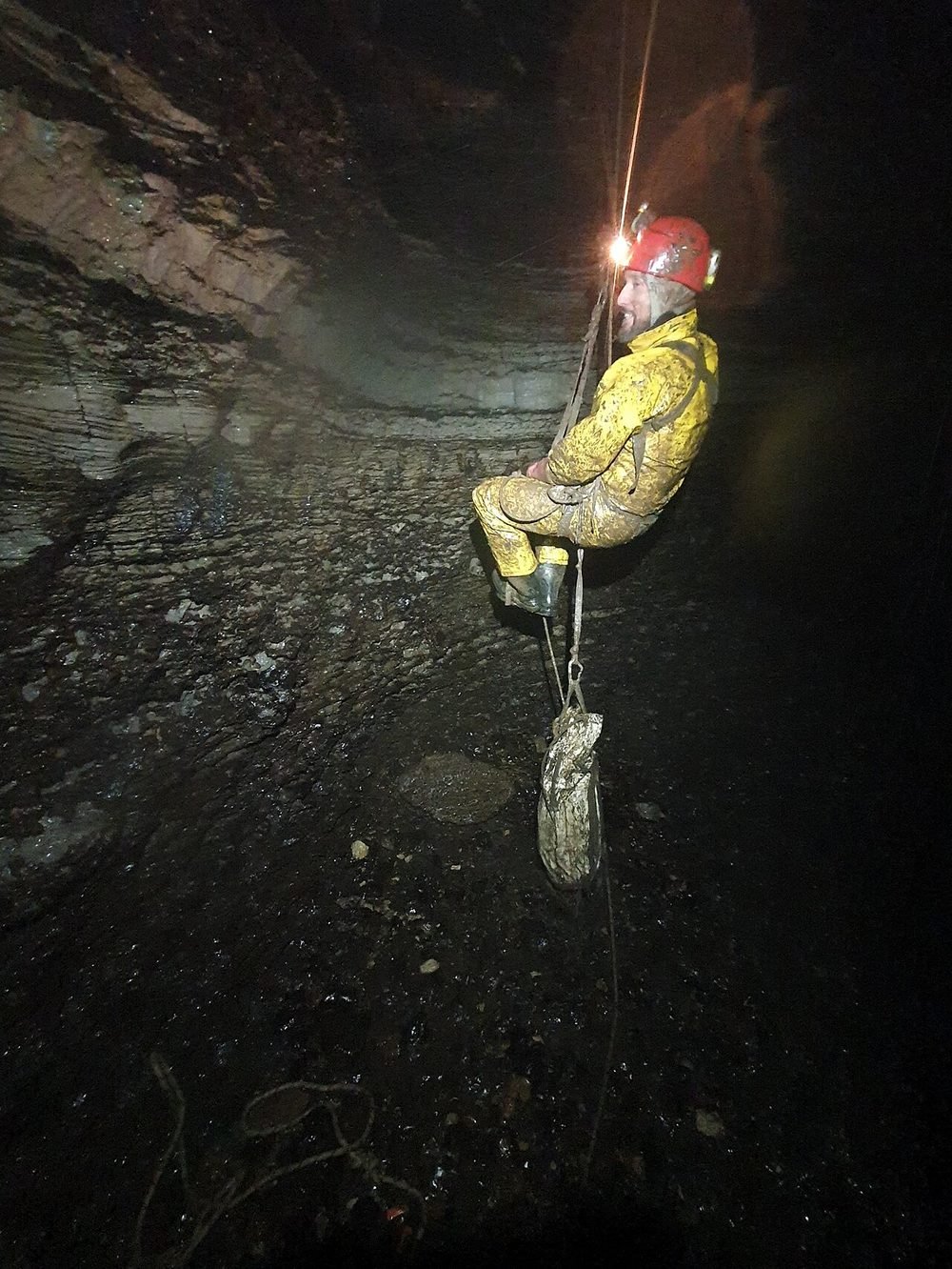 A caver hanging on a rope inside a dark cave, lit by a headlamp
