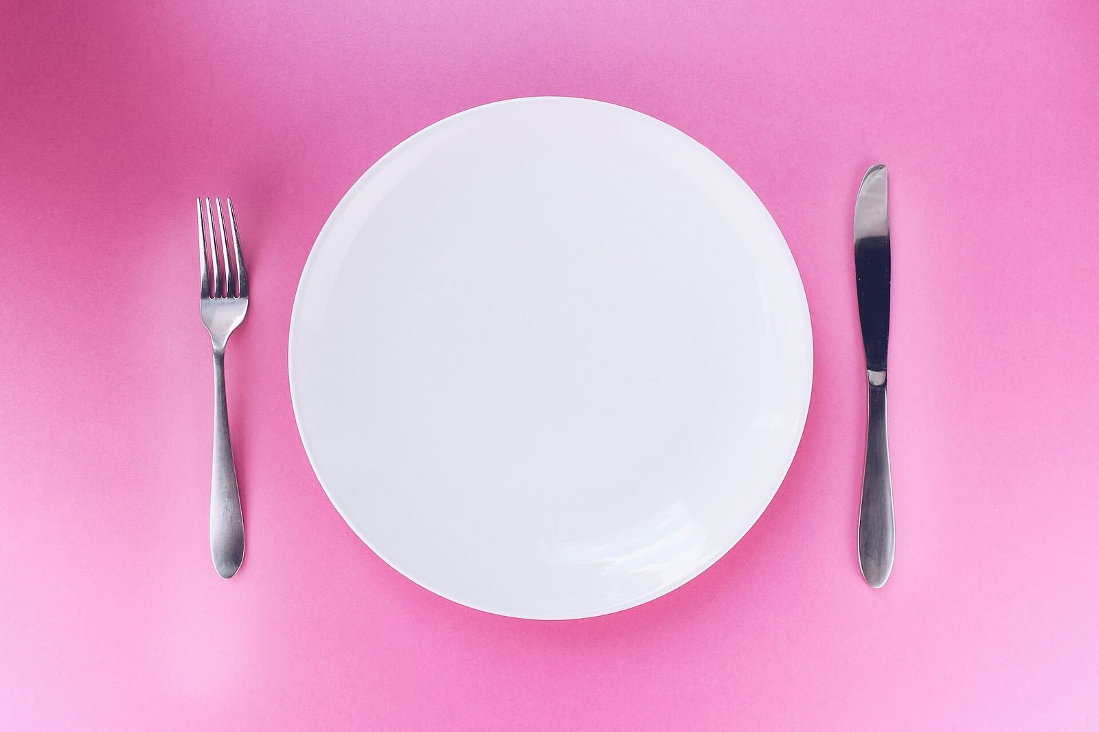 An empty white plate with a knife and fork on a pink table