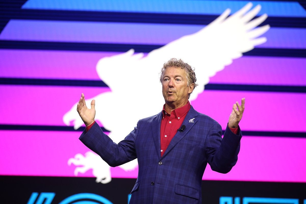 Senator Rand Paul in a blue jacket and red shirt speaking on stage with both hands raised
