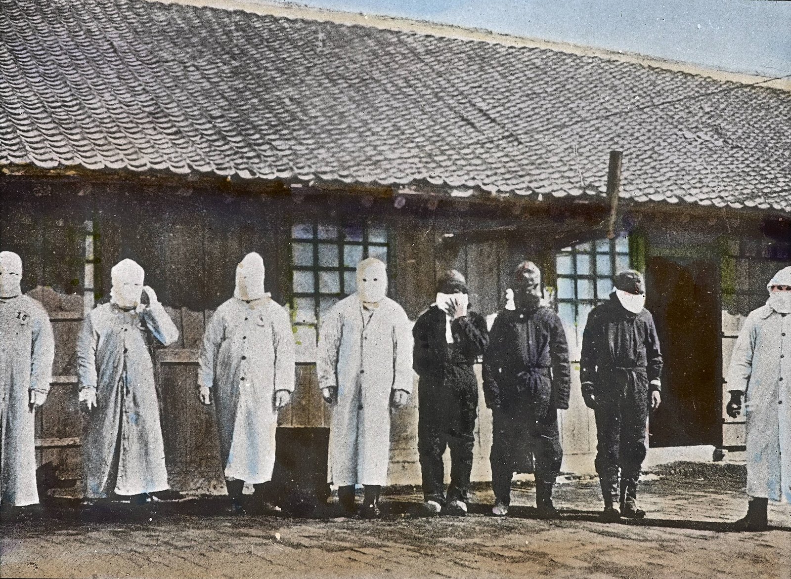 Plague workers in long white coats and cloth face masks standing outside a wooden hospital in Mukden, Manchuria, in 1911